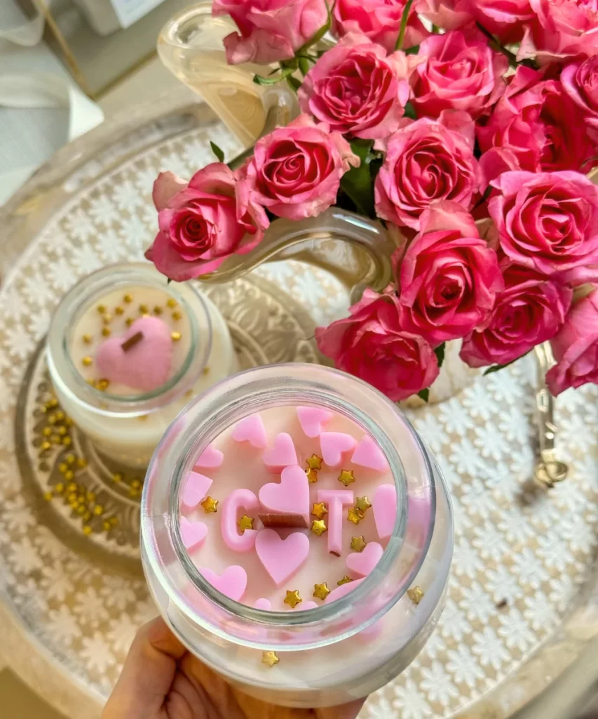 A glass jar candle made from natural soy wax with a custom name label, softly lit and surrounded by cozy décor elements.