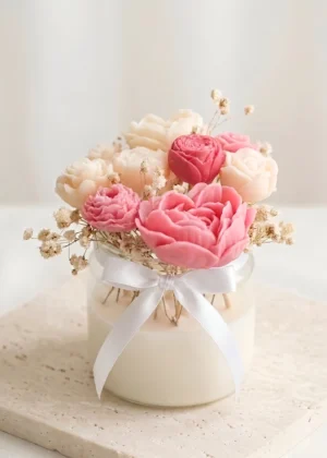 Handmade peony and rose shaped candle bouquet in blush pink and ivory wax with dried baby's breath and white satin ribbon bow on a textured stone surface